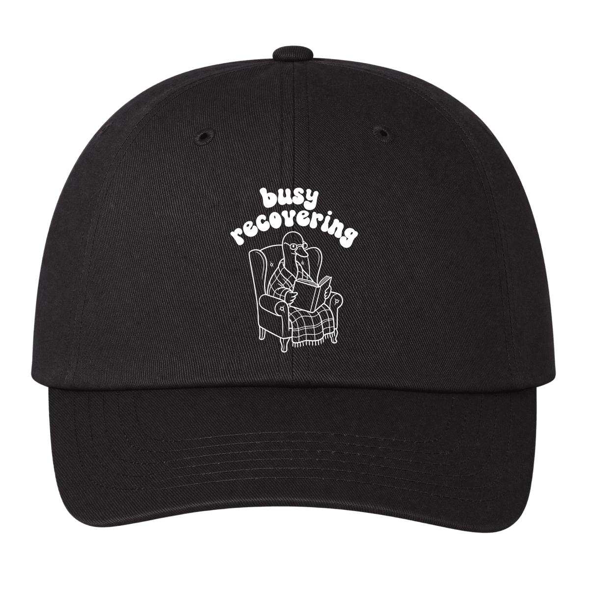 A black baseball cap with a white embroidered graphic of a goose reading a book in an armchair, with the text 'busy recovering' above it, shown against a plain white background.