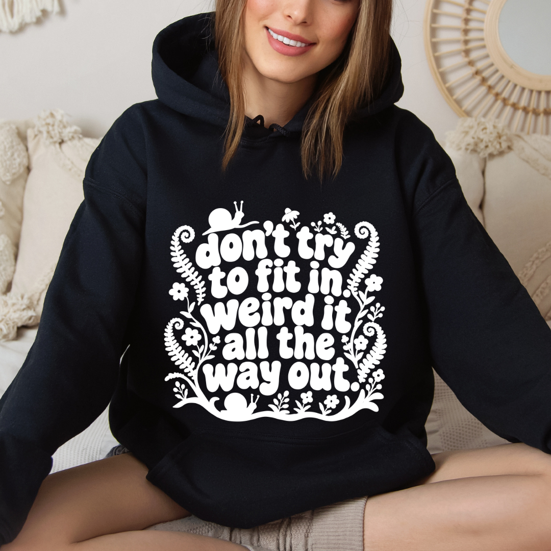 A woman wearing a black pullover hoodie with a white graphic text print that reads 'don't try to fit in weird it all the way out.' She is sitting on a beige couch in a cozy indoor setting.
