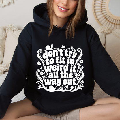 A woman wearing a black pullover hoodie with a white graphic text print that reads 'don't try to fit in weird it all the way out.' She is sitting on a beige couch in a cozy indoor setting.