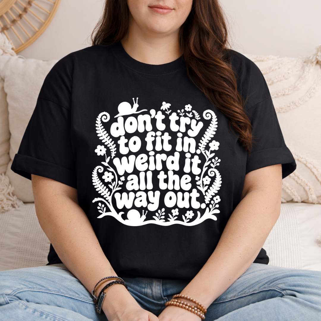 A woman wearing a black graphic t-shirt with a white hand-drawn style text design that reads 'don't try to fit in weird it all the way out.' She is sitting on a beige couch with neutral-toned pillows.