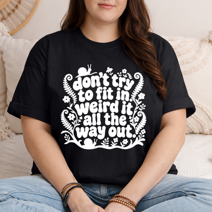 A woman wearing a black graphic t-shirt with a white hand-drawn style text design that reads 'don't try to fit in weird it all the way out.' She is sitting on a beige couch with neutral-toned pillows.