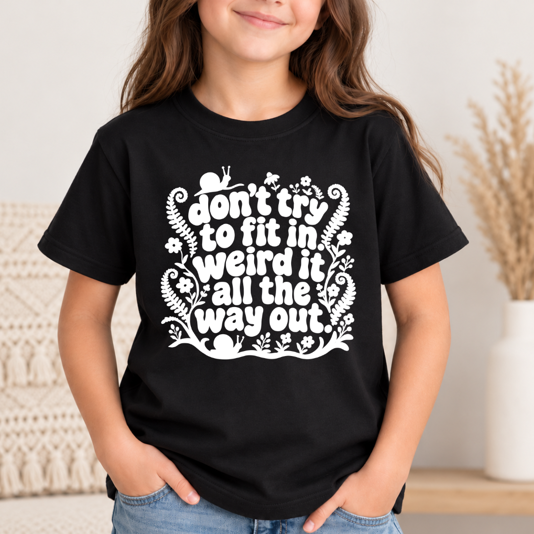 A young girl wearing a black short-sleeve graphic t-shirt with a white printed quote that reads 'don't try to fit in weird it all the way out' surrounded by cute cartoon animals and floral designs. She is standing indoors with her hands in her jeans pockets.