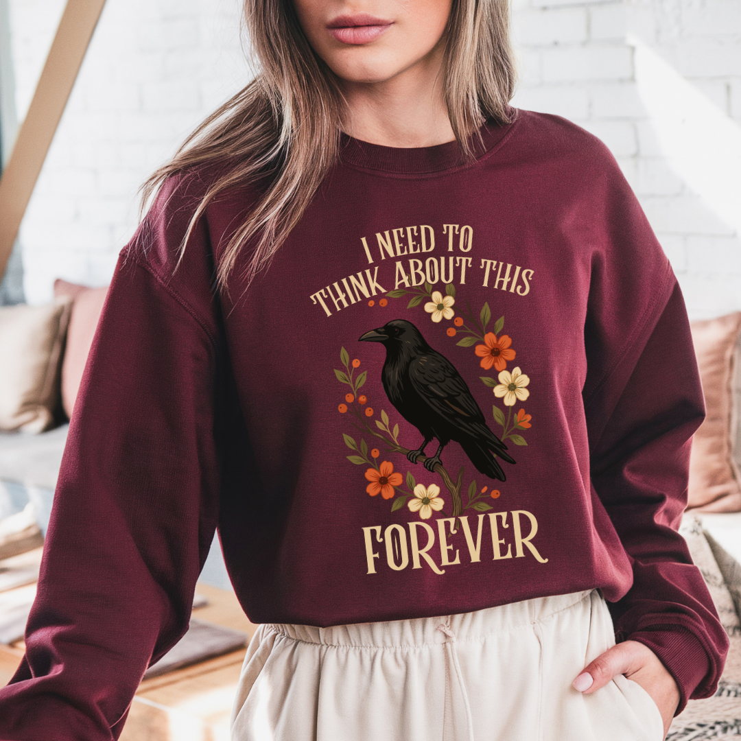 A woman wearing a maroon crewneck sweatshirt with a graphic of a black raven surrounded by orange and cream flowers, with the text 'I NEED TO THINK ABOUT THIS FOREVER' in cream lettering.