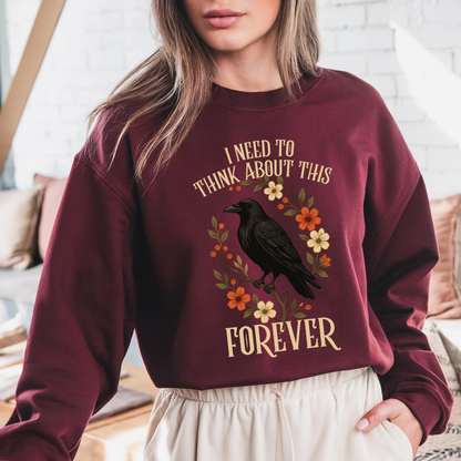 A woman wearing a maroon crewneck sweatshirt with a graphic of a black raven surrounded by orange and cream flowers, with the text 'I NEED TO THINK ABOUT THIS FOREVER' in cream lettering.