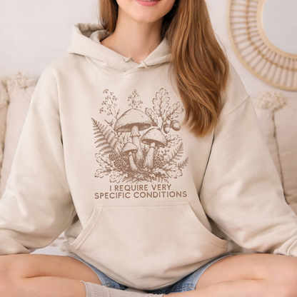 A woman wearing a beige hoodie with a brown graphic of mushrooms, ferns, and oak leaves, with the text 'I REQUIRE VERY SPECIFIC CONDITIONS' printed below. She is sitting on a beige couch.
