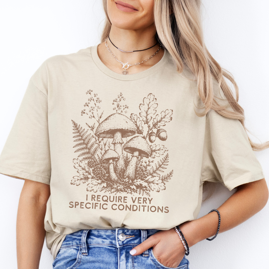 A woman wearing a beige graphic t-shirt with a brown forest mushroom design and the text 'I REQUIRE VERY SPECIFIC CONDITIONS'.