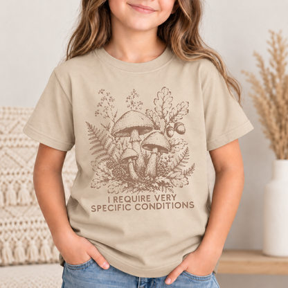 A child wearing a sand graphic t-shirt with a brown mushroom illustration and the text 'I REQUIRE VERY SPECIFIC CONDITIONS'.