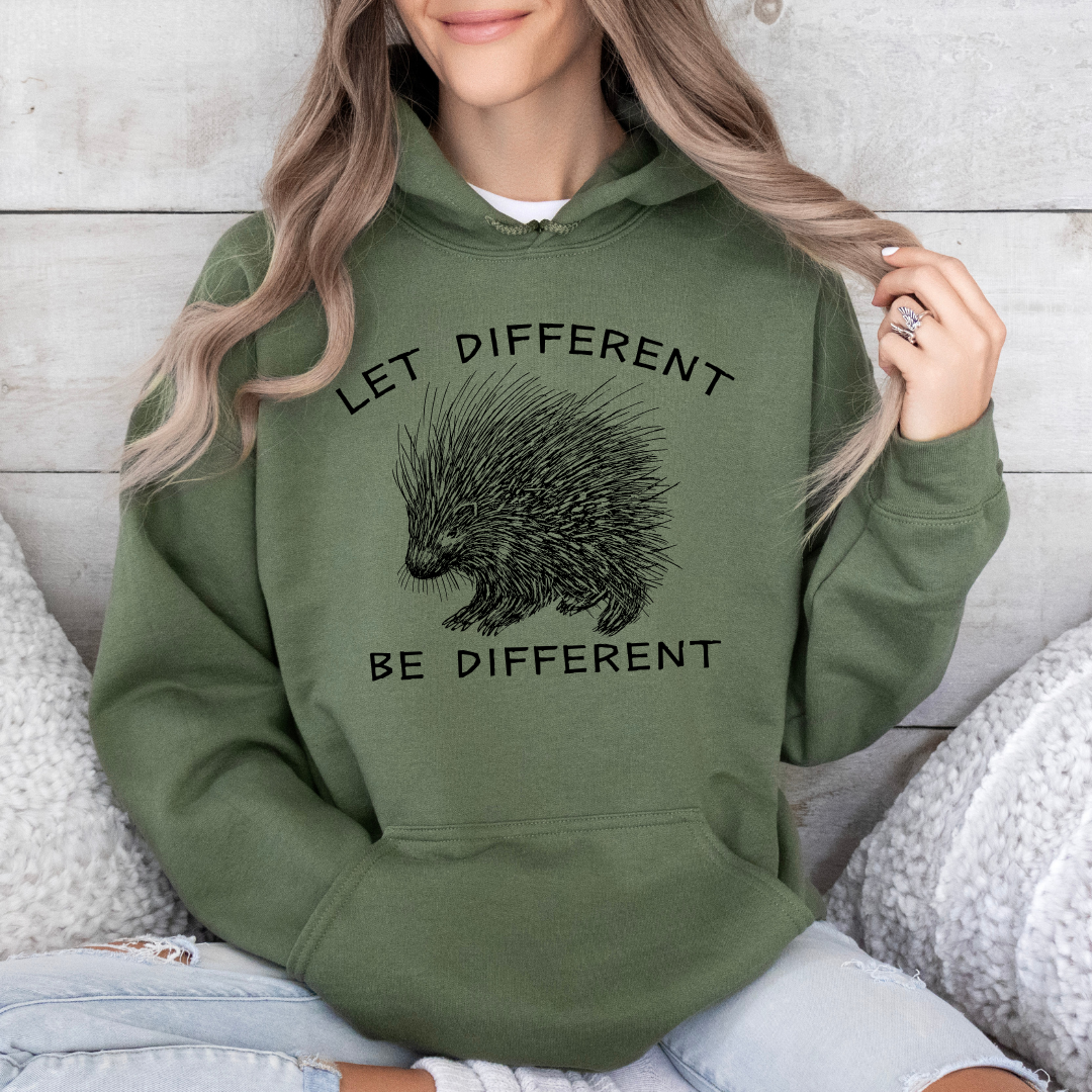 A woman wearing a military green hoodie with a black graphic of a porcupine and the text 'LET DIFFERENT BE DIFFERENT', sitting against a light wood wall.
