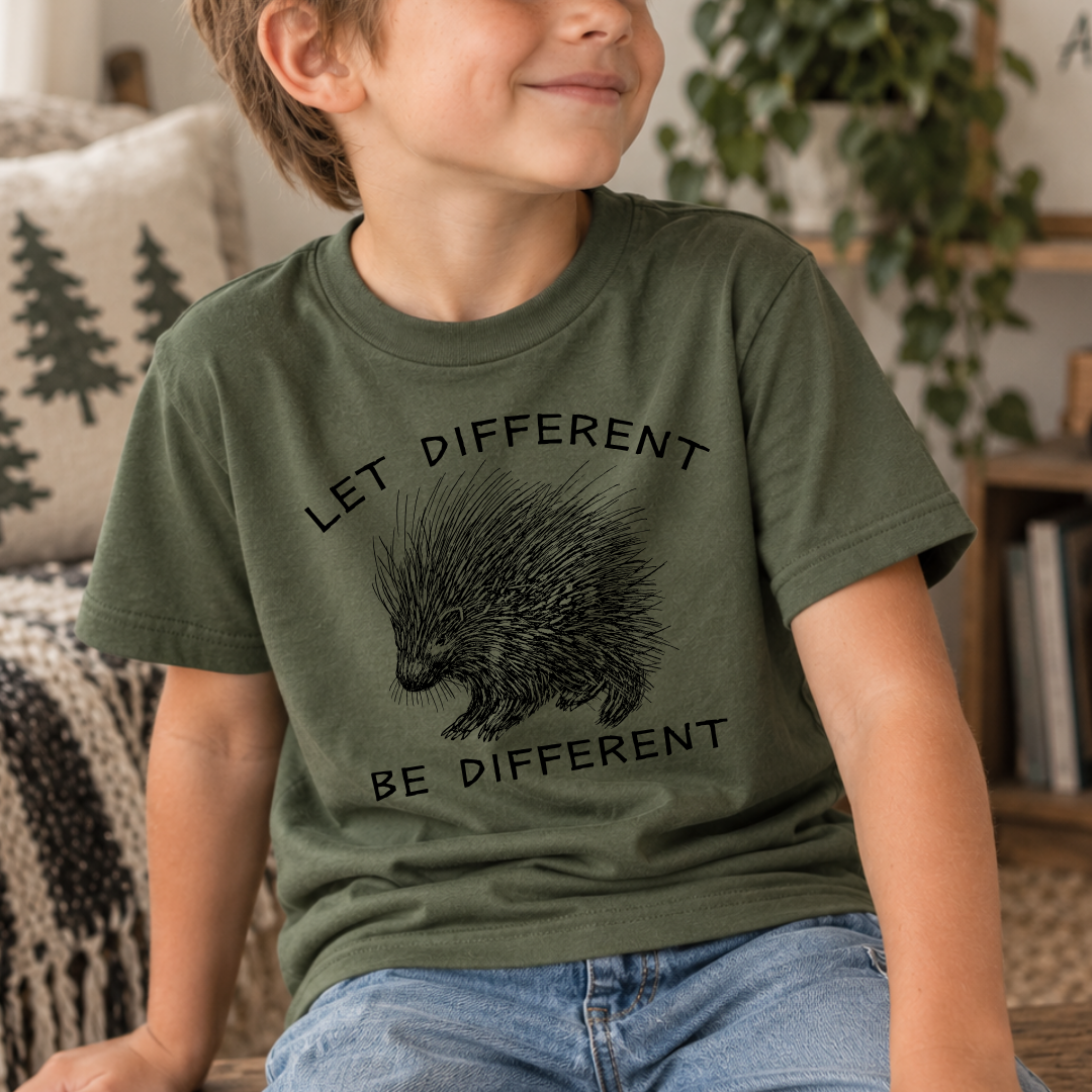 A child wearing a military green graphic t-shirt with a black printed hedgehog and the text 'LET DIFFERENT BE DIFFERENT', paired with blue jeans in a cozy indoor setting.