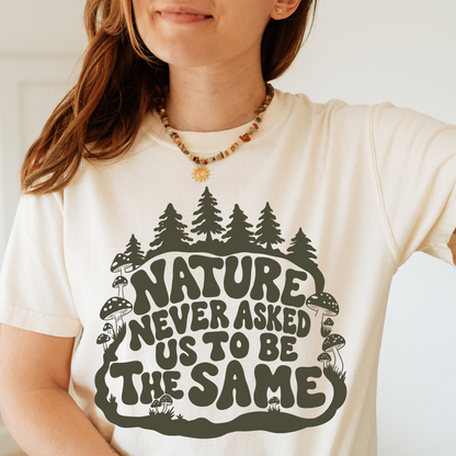 A woman wearing a cream-colored t-shirt with a dark green graphic that reads 'NATURE NEVER ASKED US TO BE THE SAME' surrounded by pine trees, mushrooms, and a deer. 