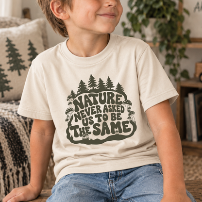 A child wearing a cream-colored t-shirt with a dark green forest graphic and the text 'NATURE NEVER ASKED US TO BE THE SAME', paired with blue jeans in a cozy indoor setting.