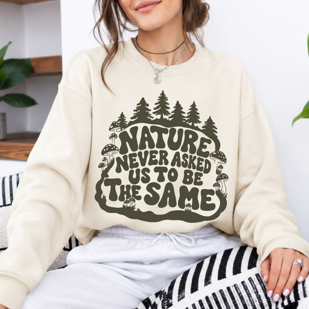 A woman wearing a cream-colored crewneck sweatshirt with a dark olive green graphic that reads 'NATURE NEVER ASKED US TO BE THE SAME' surrounded by pine trees and mushrooms, paired with white sweatpants.