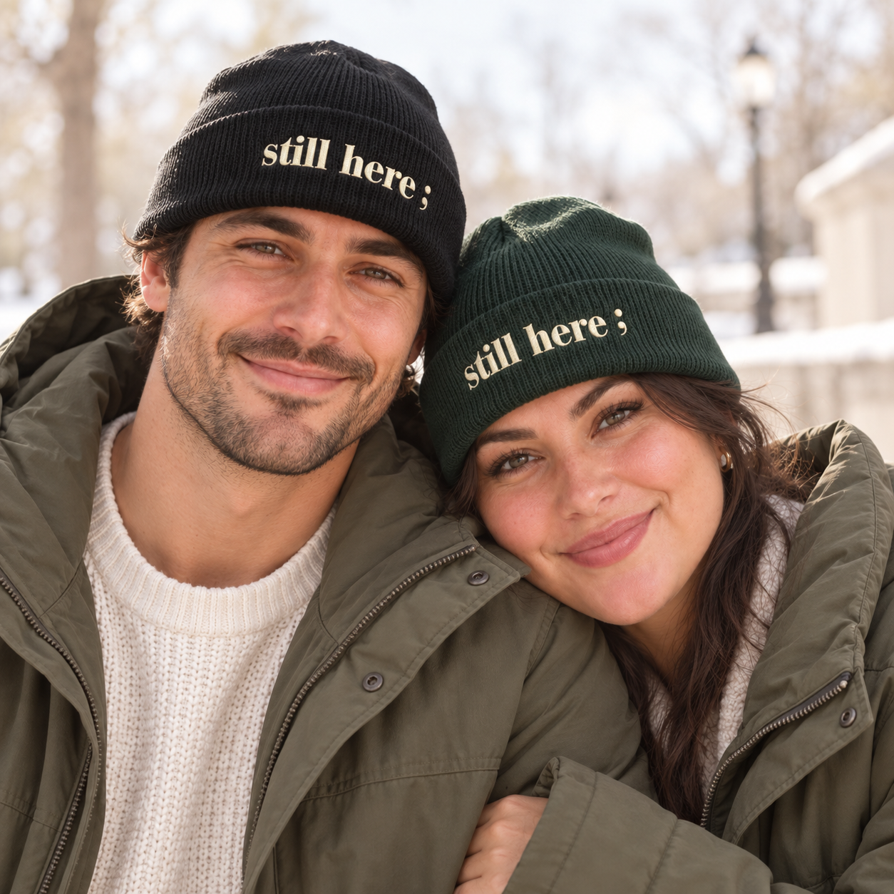 A man and a woman wearing knit beanie hats with the text 'still here ;' in white. The man's hat is black and the woman's is dark green, both photographed outdoors in a snowy setting.