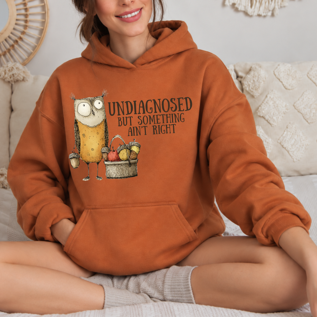 A woman sitting cross-legged on a bed wearing a rust-orange hoodie with a graphic of an owl and the text 'UNDIAGNOSED BUT SOMETHING AIN'T RIGHT'.