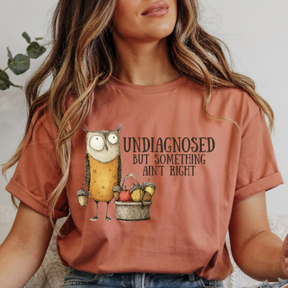 A woman wearing a rust-orange graphic t-shirt with a cartoon owl and the text 'UNDIAGNOSED BUT SOMETHING AIN'T RIGHT'.