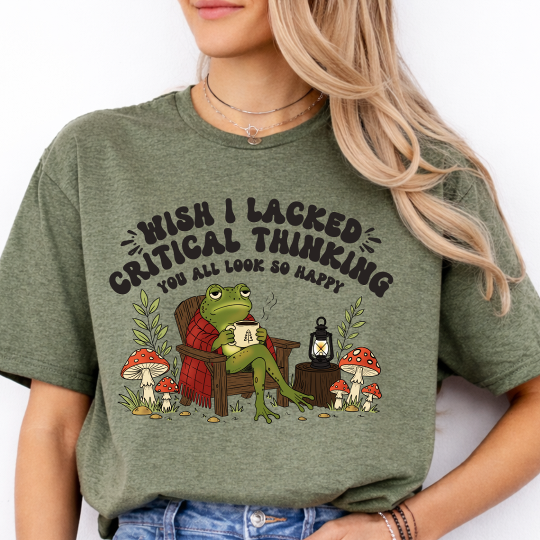 A woman wearing a military  green graphic t-shirt with a cartoon frog sitting in an armchair surrounded by mushrooms and the text 'WISH I LACKED CRITICAL THINKING YOU ALL LOOK SO HAPPY'.