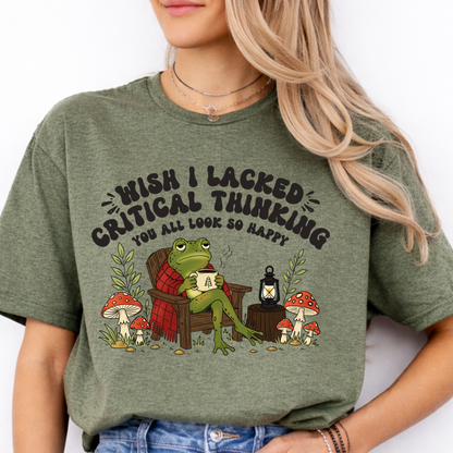 A woman wearing a military  green graphic t-shirt with a cartoon frog sitting in an armchair surrounded by mushrooms and the text 'WISH I LACKED CRITICAL THINKING YOU ALL LOOK SO HAPPY'.
