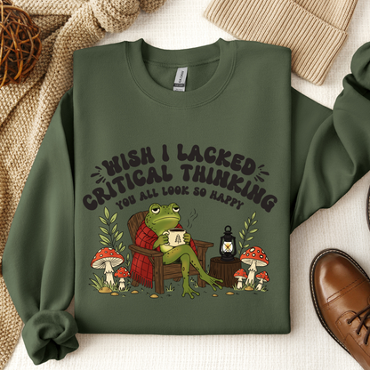 A military green crewneck sweatshirt with a humorous graphic of a green frog sitting in a chair reading a book, surrounded by mushrooms and a lantern, with the text 'WISH I LACKED CRITICAL THINKING YOU ALL LOOK SO HAPPY'. The sweatshirt is displayed in a flat-lay arrangement with a knit blanket, a brown leather shoe, and a woven ball on a light fabric background.