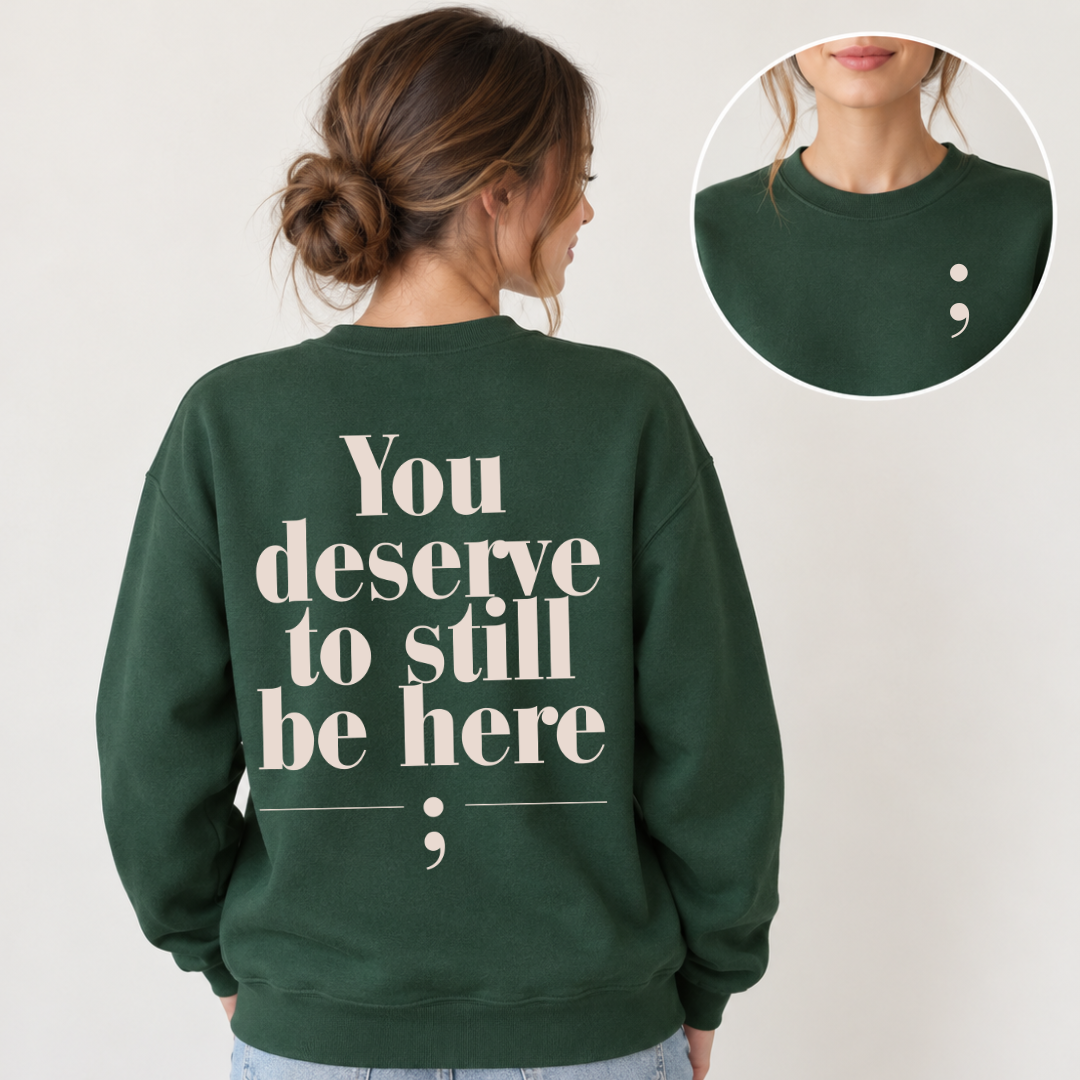 A woman shown from the back wearing a dark green crewneck sweatshirt with the phrase 'You deserve to still be here' printed in large cream serif text, plus a semicolon graphic. A circular inset at the top right shows a close-up of the back collar area with a small semicolon detail.