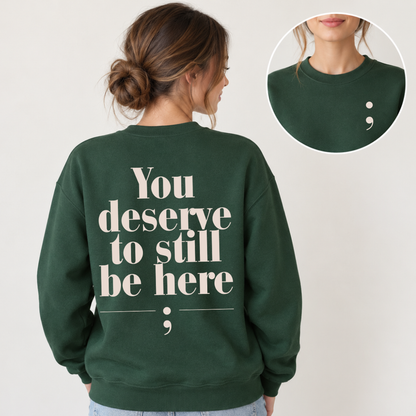 A woman shown from the back wearing a dark green crewneck sweatshirt with the phrase 'You deserve to still be here' printed in large cream serif text, plus a semicolon graphic. A circular inset at the top right shows a close-up of the back collar area with a small semicolon detail.