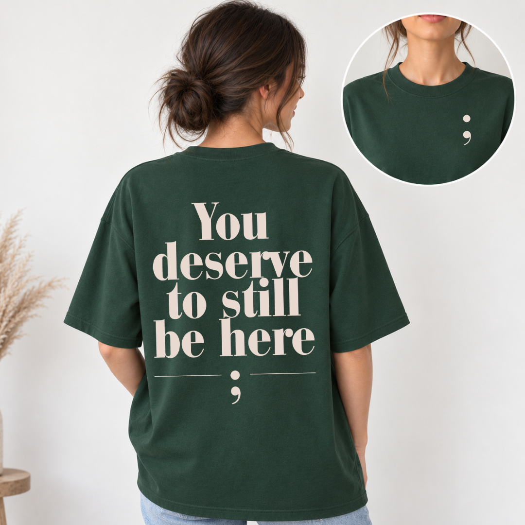 A woman wearing an oversized dark green t-shirt with the text 'You deserve to still be here' printed in cream on the back, shown from behind. A circular inset in the top right shows the front of the t-shirt with a small semicolon graphic.