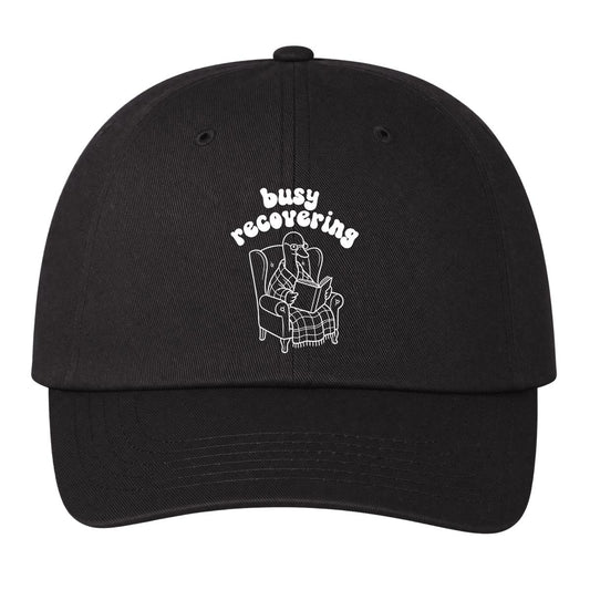 A black baseball cap with a white embroidered graphic of a goose reading a book in an armchair, with the text 'busy recovering' above it, shown against a plain white background.