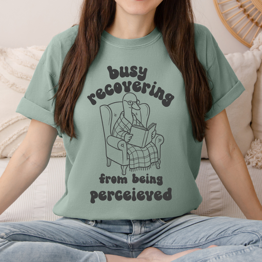 A woman wearing a sage green graphic t-shirt with a cartoon illustration of a goose  reading in an armchair. The text on the shirt reads 'busy recovering from being perceived'.