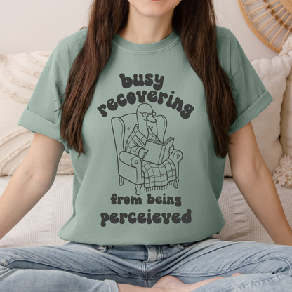 A woman wearing a sage green graphic t-shirt with a cartoon illustration of a goose  reading in an armchair. The text on the shirt reads 'busy recovering from being perceived'.
