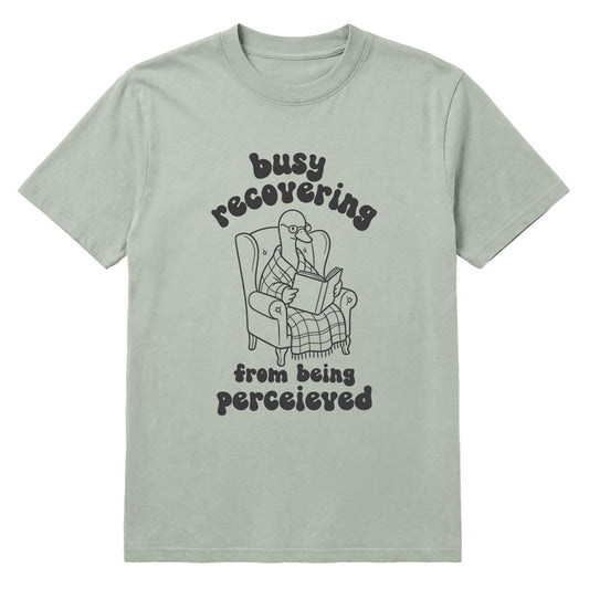 A sage green crewneck t-shirt featuring a charcoal graphic of a goose reading a newspaper in an armchair, with the text 'busy recovering from being perceived' written around the illustration.