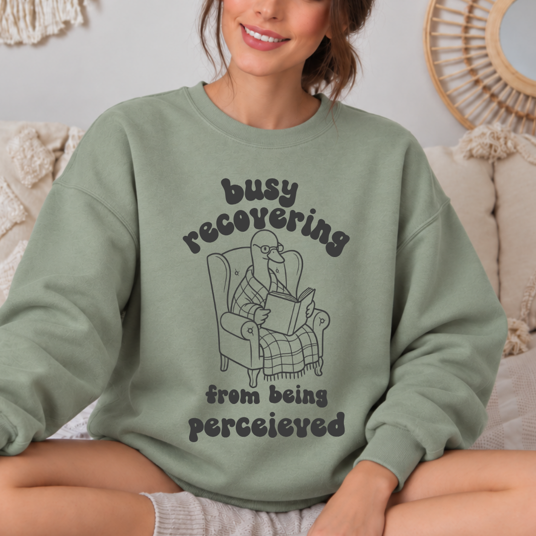 A woman wearing a sage green crewneck sweatshirt with a black graphic of a goose reading in an armchair and the text 'busy recovering from being perceieved'.