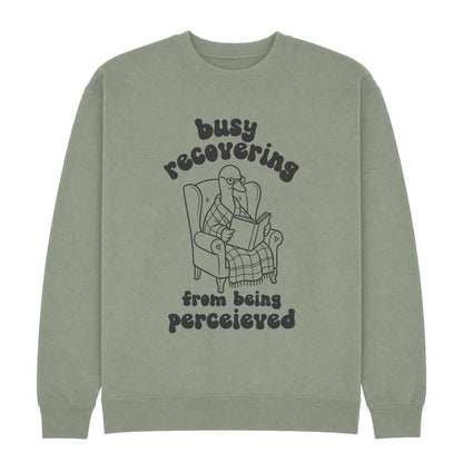 A sage green crewneck sweatshirt featuring a dark gray graphic of a goose reading in an armchair with the text 'busy recovering from being perceived'.