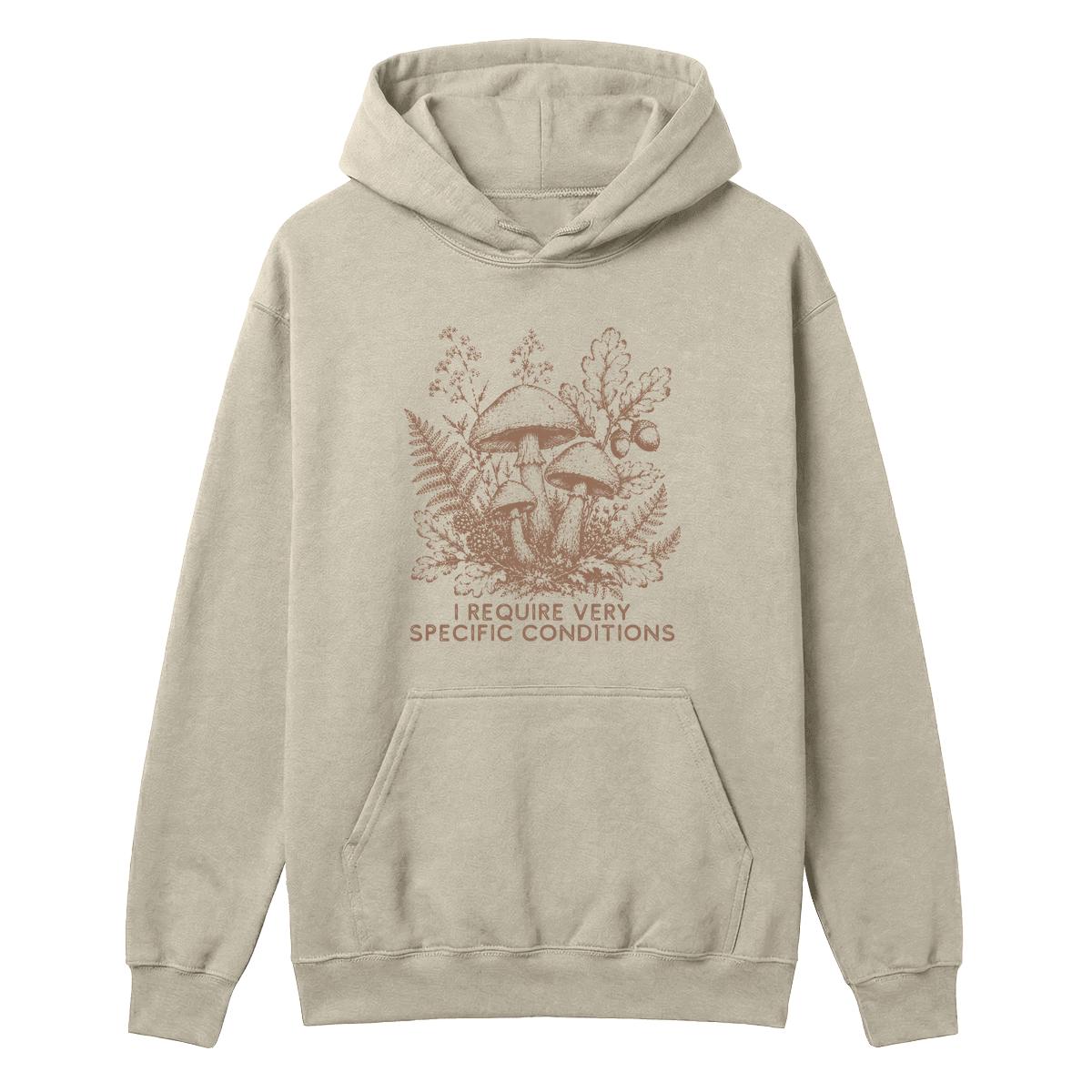 A light beige pullover hoodie with a brown graphic of mushrooms, ferns, and the text 'I REQUIRE VERY SPECIFIC CONDITIONS' on the front, displayed against a solid white background.
