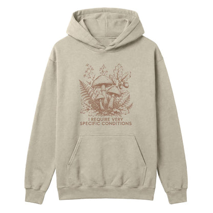 A light beige pullover hoodie with a brown graphic of mushrooms, ferns, and the text 'I REQUIRE VERY SPECIFIC CONDITIONS' on the front, displayed against a solid white background.
