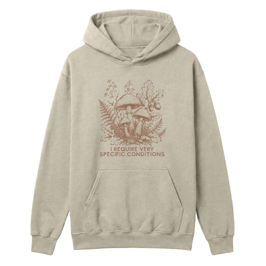 A light beige pullover hoodie with a brown graphic of mushrooms, ferns, and the text 'I REQUIRE VERY SPECIFIC CONDITIONS' on the front, displayed against a solid white background.
