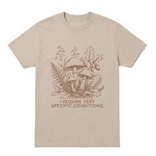 A beige crewneck t-shirt with a brown graphic of mushrooms, ferns, and oak leaves, with the text 'I REQUIRE VERY SPECIFIC CONDITIONS' printed below.