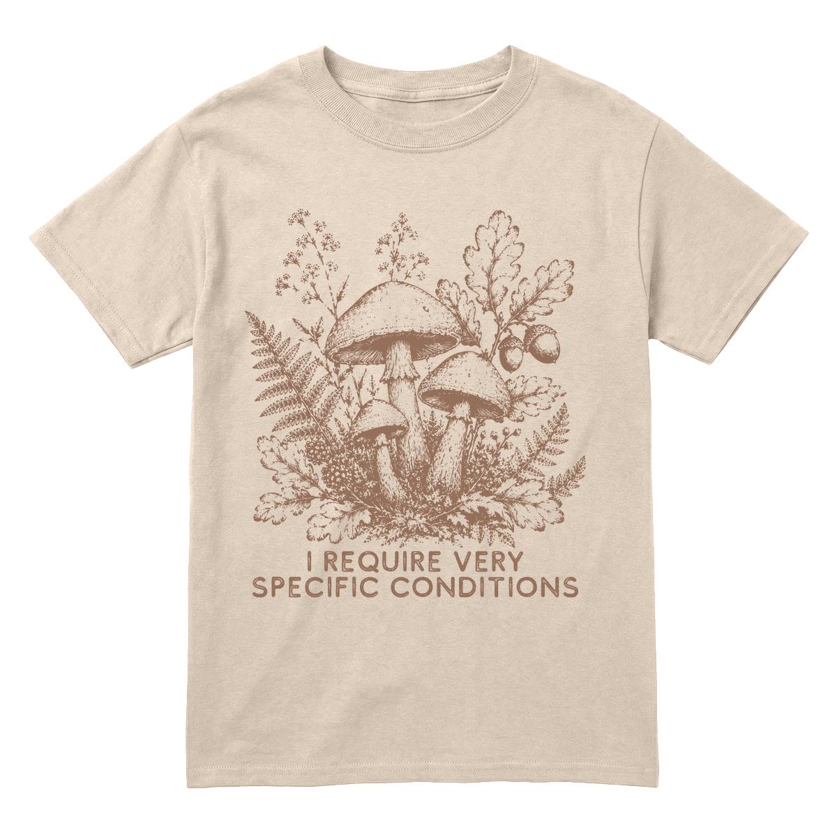 A sand crewneck t-shirt featuring a brown line art graphic of mushrooms, ferns, and oak leaves, with the text 'I REQUIRE VERY SPECIFIC CONDITIONS' printed below.