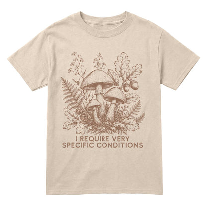 A sand crewneck t-shirt featuring a brown line art graphic of mushrooms, ferns, and oak leaves, with the text 'I REQUIRE VERY SPECIFIC CONDITIONS' printed below.