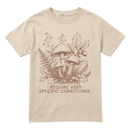A sand crewneck t-shirt featuring a brown line art graphic of mushrooms, ferns, and oak leaves, with the text 'I REQUIRE VERY SPECIFIC CONDITIONS' printed below.