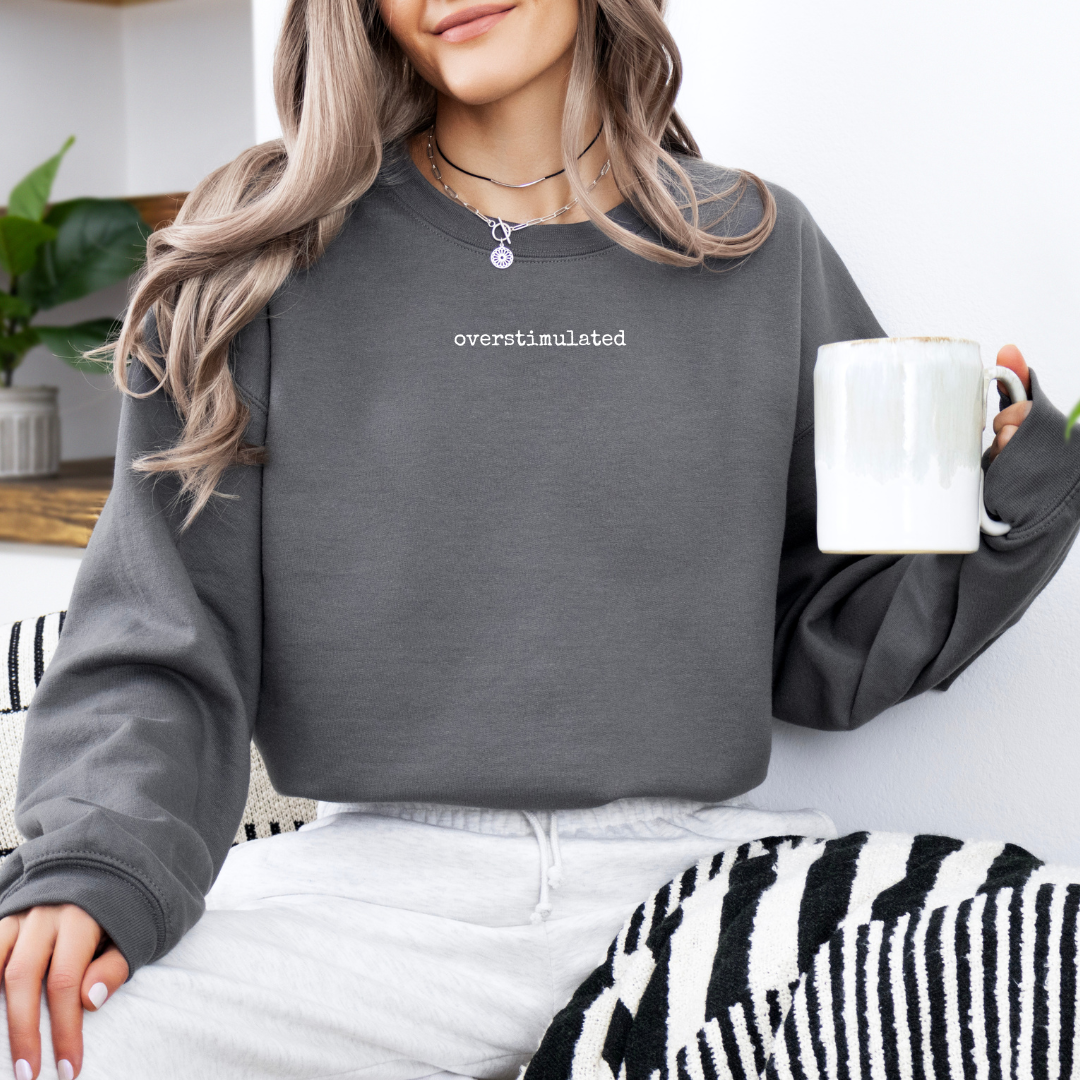 Woman wearing a charcoal “overstimulated” crewneck sweatshirt while seated indoors holding a white mug.