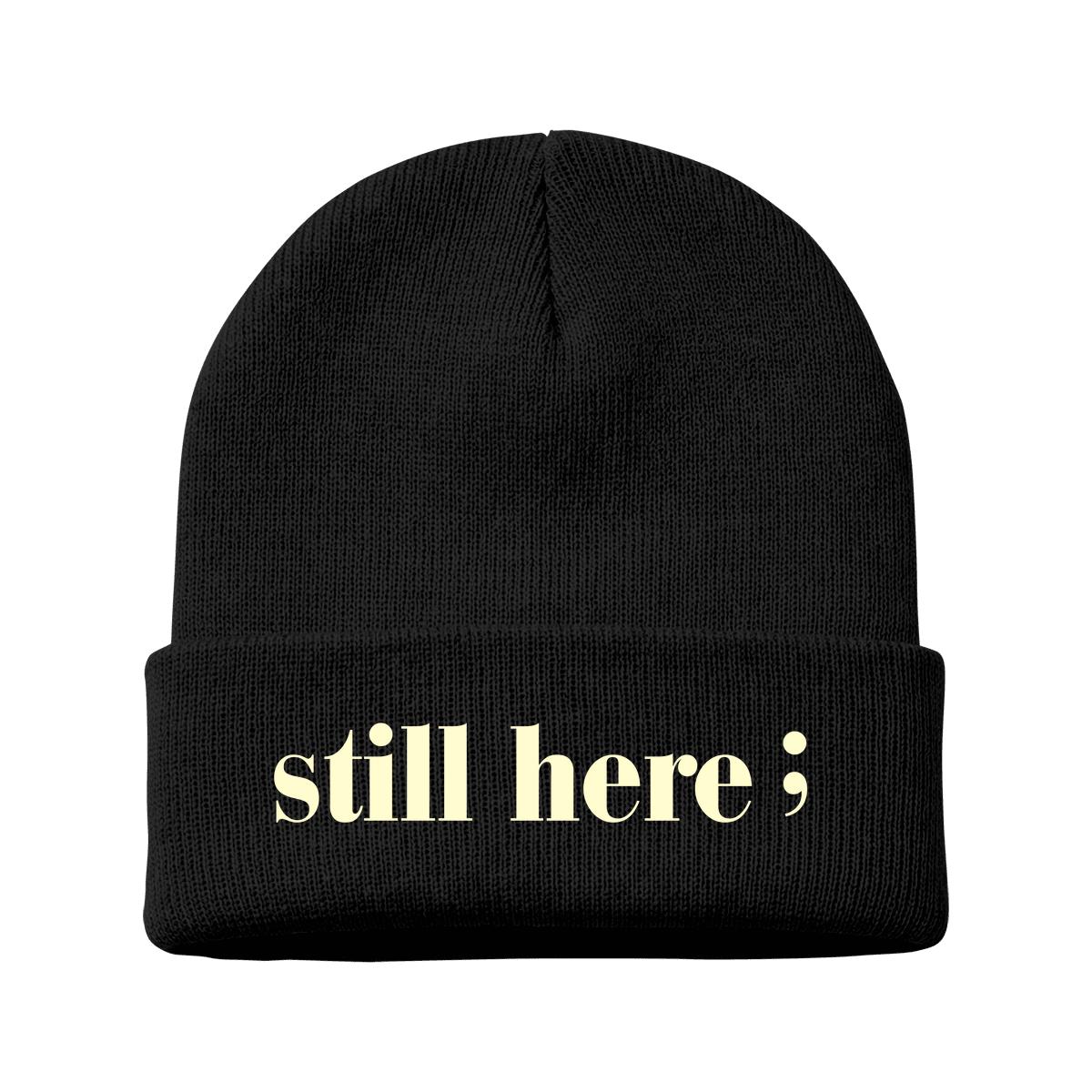 A black knit beanie with a folded cuff featuring the text 'still here;' in a pale cream serif font, shown against a plain white background.