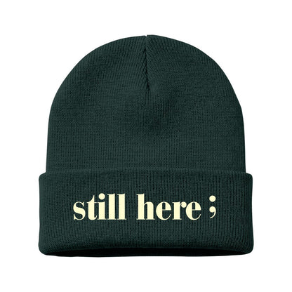 A dark forest green ribbed knit beanie with a folded cuff, featuring the text 'still here;' in a pale yellow serif font on the front, set against a plain white background.