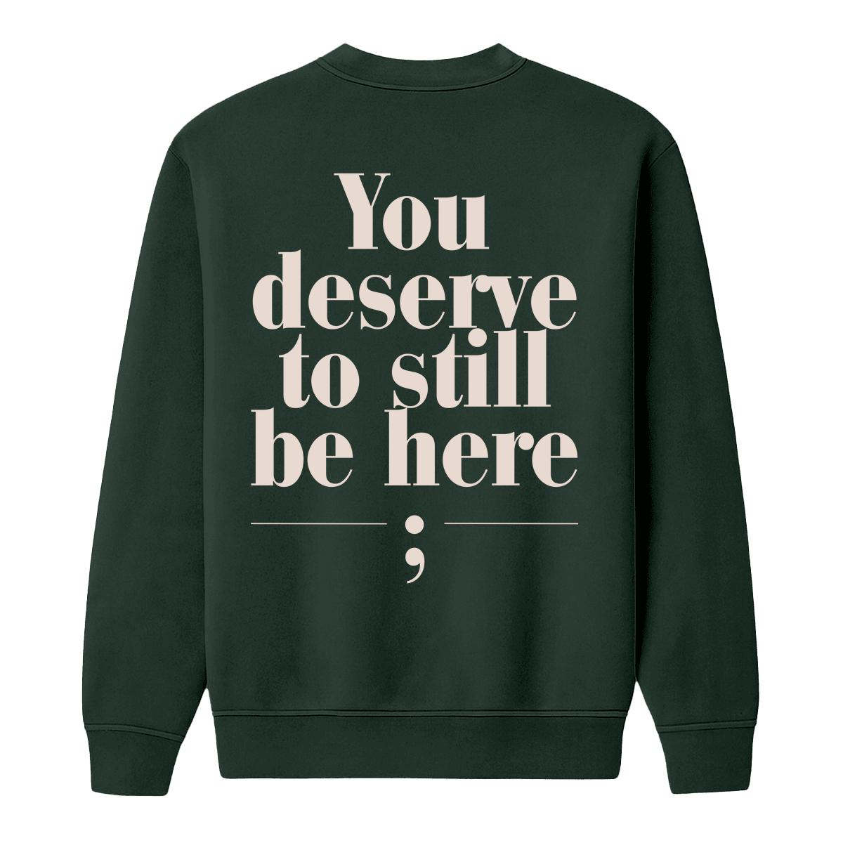 A dark forest green crewneck sweatshirt shown from the back, featuring the text 'You deserve to still be here;' printed in a large, cream serif font with a semicolon symbol underneath.