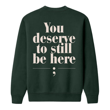A dark forest green crewneck sweatshirt shown from the back, featuring the text 'You deserve to still be here;' printed in a large, cream serif font with a semicolon symbol underneath.