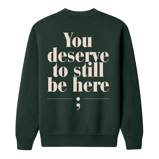 A dark forest green crewneck sweatshirt shown from the back, featuring the text 'You deserve to still be here;' printed in a large, cream serif font with a semicolon symbol underneath.