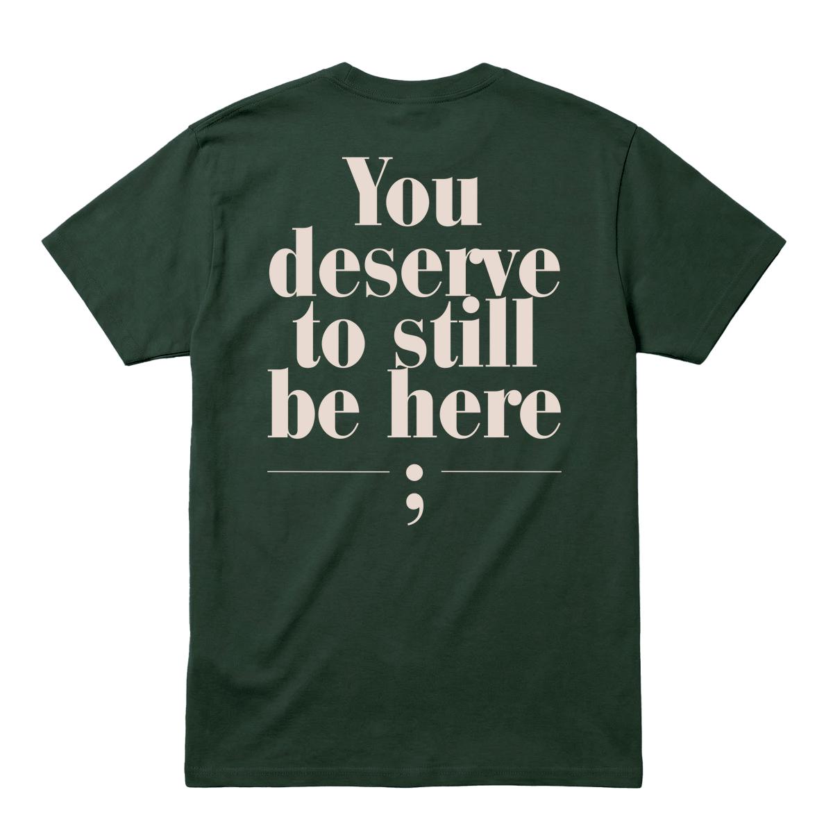A dark forest green short-sleeve t-shirt shown from the back, featuring a large cream-colored typographic print that reads 'You deserve to still be here;' on a solid white background.