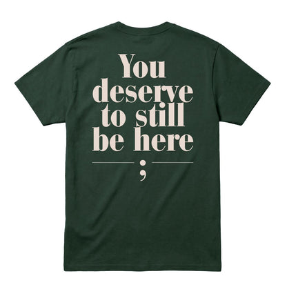 A dark forest green short-sleeve t-shirt shown from the back, featuring a large cream-colored typographic print that reads 'You deserve to still be here;' on a solid white background.
