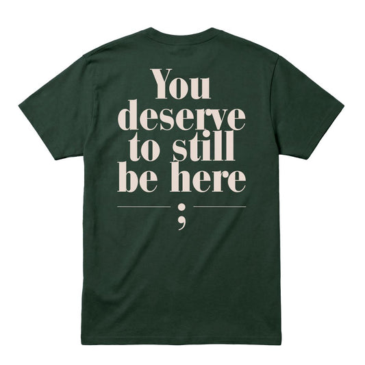 A dark forest green short-sleeve t-shirt shown from the back, featuring a large cream-colored typographic print that reads 'You deserve to still be here;' on a solid white background.